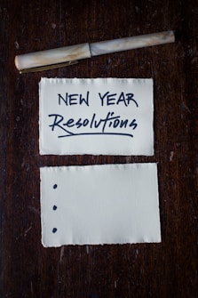 Two pieces of torn paper rest on a dark wooden surface. The top paper has 'NEW YEAR Resolutions' written in bold, black lettering. A pen is placed above the papers. The bottom paper has three bullet points but is otherwise blank.