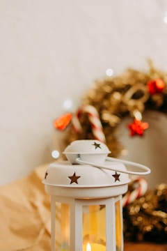 A decorative white lantern with star cutouts sits in the foreground, emitting a warm, glowing light from a candle inside. In the background, there is a festive wreath with gold tinsel, red and white candy canes, and red ornaments, placed against a soft-focus neutral-colored wall.