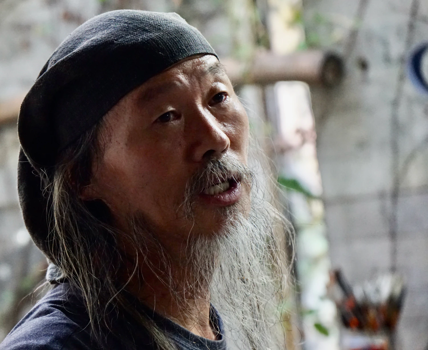 An older man in a knitted cap and a long white beard mid-sentence, a rack of brushes behind him