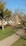 A quaint, thatched-roof cottage is set in a lush, green landscape, surrounded by trees. A large, sculptural installation made of woven natural materials is prominently displayed in front of the house. The clear blue sky adds to the serene and picturesque setting.