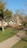 A quaint, thatched-roof cottage is set in a lush, green landscape, surrounded by trees. A large, sculptural installation made of woven natural materials is prominently displayed in front of the house. The clear blue sky adds to the serene and picturesque setting.
