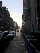 A city street scene at sunset with two rows of buildings lining a sloped road. Cars are parked along the street, and a few pedestrians walk on the sidewalk. A 'TO LET' sign hangs on one of the buildings. The sun is low in the sky, casting long shadows.