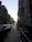 A city street scene at sunset with two rows of buildings lining a sloped road. Cars are parked along the street, and a few pedestrians walk on the sidewalk. A 'TO LET' sign hangs on one of the buildings. The sun is low in the sky, casting long shadows.