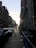 A city street scene at sunset with two rows of buildings lining a sloped road. Cars are parked along the street, and a few pedestrians walk on the sidewalk. A 'TO LET' sign hangs on one of the buildings. The sun is low in the sky, casting long shadows.