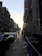 A city street scene at sunset with two rows of buildings lining a sloped road. Cars are parked along the street, and a few pedestrians walk on the sidewalk. A 'TO LET' sign hangs on one of the buildings. The sun is low in the sky, casting long shadows.