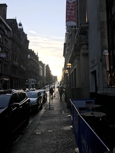 A city street scene at sunset with two rows of buildings lining a sloped road. Cars are parked along the street, and a few pedestrians walk on the sidewalk. A 'TO LET' sign hangs on one of the buildings. The sun is low in the sky, casting long shadows.