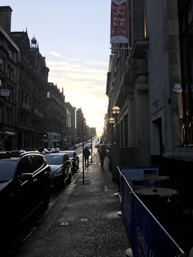 A city street scene at sunset with two rows of buildings lining a sloped road. Cars are parked along the street, and a few pedestrians walk on the sidewalk. A 'TO LET' sign hangs on one of the buildings. The sun is low in the sky, casting long shadows.