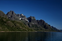 A stunning landscape of tall, rugged mountains with patches of snow on their peaks, surrounded by lush greenery. The mountains reflect perfectly in the calm water of a fjord under a clear blue sky.