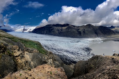 A vast glacier flows down from a rugged mountain range under a bright blue sky with scattered clouds. The foreground consists of rocky terrain with patches of green vegetation. The glacier is streaked with dark lines, likely indicating crevasses or debris. In the distance, icebergs float on a glacial lake.