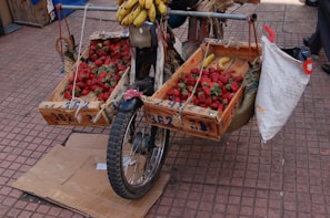 A makeshift fruit cart is attached to the front of a motorbike, displaying wooden crates filled with strawberries and bananas. The cart is positioned on a tiled floor with some cardboard underneath the front wheel.