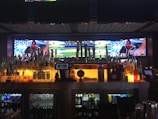 A bar with a wide selection of liquor bottles and beer taps is in the foreground, with a digital screen displaying a baseball game in the background. The screen shows multiple angles of the game, including players and the field. The bar is dimly lit with an amber glow beneath the bottles and taps, creating a cozy atmosphere.