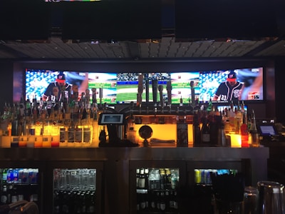 A bar with a wide selection of liquor bottles and beer taps is in the foreground, with a digital screen displaying a baseball game in the background. The screen shows multiple angles of the game, including players and the field. The bar is dimly lit with an amber glow beneath the bottles and taps, creating a cozy atmosphere.