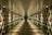A long, symmetrical corridor with reflective glass walls and a patterned ceiling, illuminated by warm artificial lights. The perspective converges towards a distant dark opening, creating a sense of depth and infinity.
