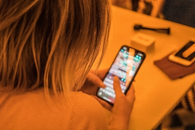 A person with blonde hair is using a smartphone, typing on a touchscreen. The phone screen is bright and displays a messaging or social media app. The background is softly blurred brown hues, with some indistinct objects nearby, like a small box and another mobile device on a table.