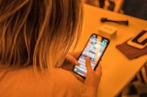 A person with blonde hair is using a smartphone, typing on a touchscreen. The phone screen is bright and displays a messaging or social media app. The background is softly blurred brown hues, with some indistinct objects nearby, like a small box and another mobile device on a table.