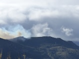 A mountainous landscape under a cloudy sky, with a plume of smoke rising from behind one of the hills. The hills are covered with green vegetation, and the clouds appear thick and gray, suggesting an overcast atmosphere.