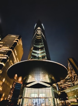 A modern skyscraper with a sleek, curved design dominates the scene. The building is illuminated, casting a warm glow against the night sky. The entrance is grand, featuring glass doors and decorative lights. Surrounding structures are similarly lit, contributing to a cityscape filled with reflections and light.