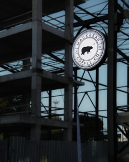 A round signboard with the text 'MACKBEAR COFFEE CO.' is hanging against the backdrop of a building under construction. The sign features the silhouette of a bear in the center. The structure behind is made of concrete and steel beams, partially enclosed, indicating ongoing construction. The sky is overcast, contributing to a subdued lighting.