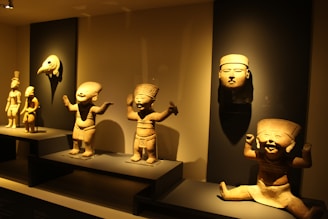 A display of ancient sculptures and artifacts in a dimly lit museum setting. The collection includes several human-like figurines with exaggerated features and a large human face mask. The pieces are arranged artistically on dark platforms to highlight their intricate designs and craftsmanship.