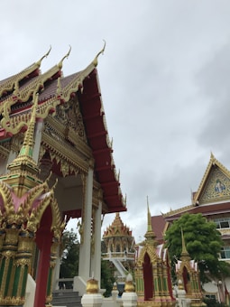 A traditional Buddhist temple with intricately decorated roofs adorned with gold and red accents. The architecture features ornate details and spires, set against a cloudy sky. Lush green trees are visible around the structures.