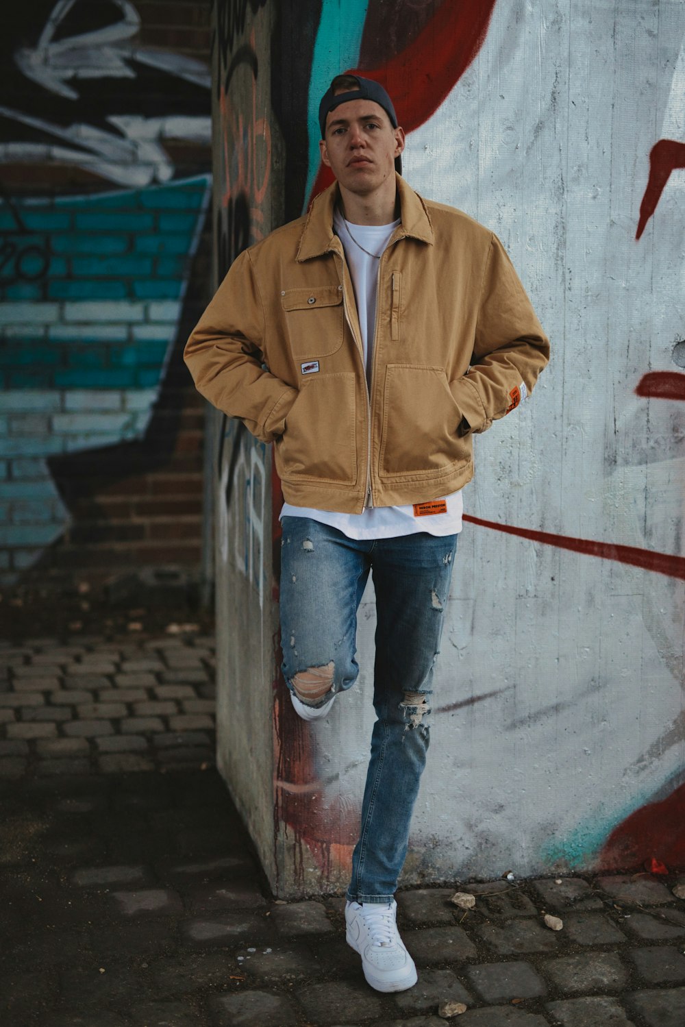man in brown leather jacket and blue denim jeans leaning on white wall