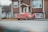 A red car featuring a number and flame decal is parked in the driveway of a brown house. The house has multiple windows and a small lawn, and it's situated in a residential area with similar homes visible nearby.