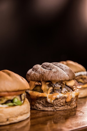 Three gourmet hamburgers placed on a wooden surface, featuring a variety of toppings such as melted cheese, onions, and bacon. The buns have a rustic appearance with visible flour dusting.