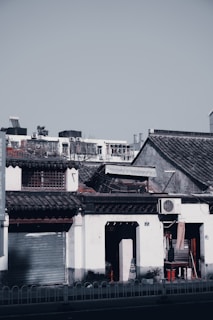 A traditional building with white walls and decorative roof tiles. The structure includes a shuttered entrance and an air conditioning unit mounted on the facade. The setting is urban, with more modern buildings visible in the background under a clear sky.