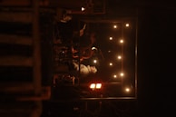 A small group of musicians are performing on a dimly lit stage. The stage is surrounded by a frame of warm string lights. The atmosphere suggests an intimate live performance in a cozy setting.