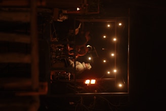 A small group of musicians are performing on a dimly lit stage. The stage is surrounded by a frame of warm string lights. The atmosphere suggests an intimate live performance in a cozy setting.