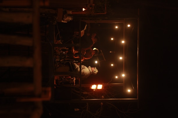 A small group of musicians are performing on a dimly lit stage. The stage is surrounded by a frame of warm string lights. The atmosphere suggests an intimate live performance in a cozy setting.