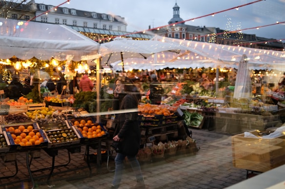 A bustling market stall under white canopies displays an array of fresh fruits and vegetables. String lights above create a warm, inviting atmosphere. People, dressed for cooler weather, walk by, interacting with the vendors. In the background, a classic European-style building adds to the charm of the scene. Reflections in a nearby glass surface provide added depth to the composition.