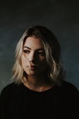 A young woman with blonde hair and subtle makeup gazes thoughtfully to the side. Her expression is calm and slightly introspective, and the soft lighting creates a dramatic shadow effect on her face. She wears a dark-colored top, and the background is a muted, blurred gradient of dark green and gray tones.