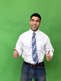 A person dressed in a white shirt and blue striped tie is standing against a green background, showing a thumbs-up gesture with both hands. The person is smiling and appears to be confident and relaxed.