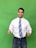A person dressed in a white shirt and blue striped tie is standing against a green background, showing a thumbs-up gesture with both hands. The person is smiling and appears to be confident and relaxed.