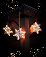 Three illuminated snowflake-shaped lights hang from a wooden structure. The background is dark with small, blurry lights scattered around, suggesting a festive or holiday setting.