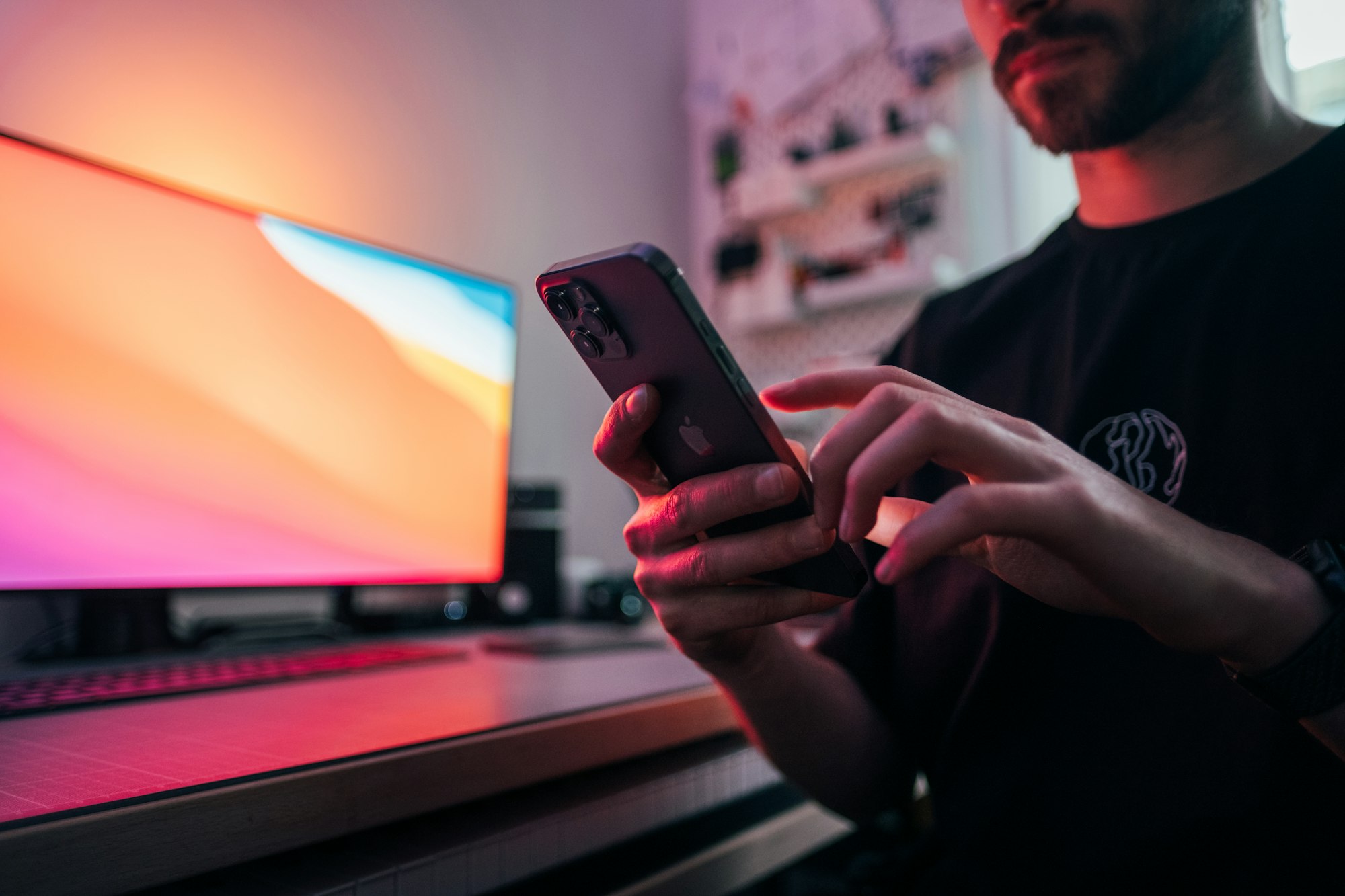 Young man holding an iPhone 12 Pro Max in front of his desk. In the background, there's a monitor using the Big Sur wallpaper. Picture taken by Jonas Leupe (www.brandstof.cc/jonas) for Tandem Tech (www.tandemtech.be)