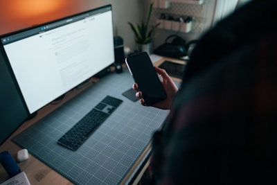 A workspace featuring a large monitor displaying a webpage, a wireless keyboard on a grid-patterned desk mat, and a hand holding a smartphone. A plant is visible in the background, along with some shelves.