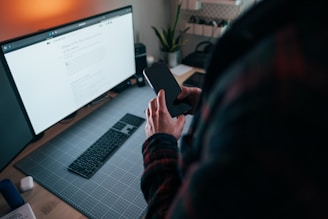 A person holds a smartphone in one hand while standing at a desk with a large computer monitor displaying a webpage. The workspace is neatly organized with a keyboard, a glowing desk lamp, and a small plant placed nearby.