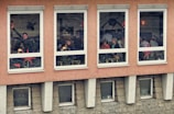 A group of people is seated in a warmly lit room inside a building, visible through four large windows. The walls are orange and decorated with star-shaped ornaments. Some of the individuals appear to be engaged in conversation or eating at tables, suggesting a cafe or restaurant setting.