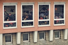 A group of people is seated in a warmly lit room inside a building, visible through four large windows. The walls are orange and decorated with star-shaped ornaments. Some of the individuals appear to be engaged in conversation or eating at tables, suggesting a cafe or restaurant setting.