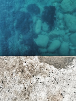 A concrete surface is shown in contrast with clear blue water. The concrete has visible small stones and pebbles embedded within, while beneath the water, large rocks are faintly visible, suggesting depth and transparency.