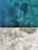 A concrete surface is shown in contrast with clear blue water. The concrete has visible small stones and pebbles embedded within, while beneath the water, large rocks are faintly visible, suggesting depth and transparency.