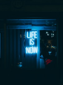 A glowing neon sign emits a bright blue light, displaying the words 'Life is Now' against a dark background. The ambient reflections and glass panels create a moody atmosphere.