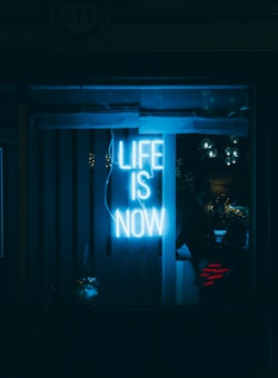 A glowing neon sign emits a bright blue light, displaying the words 'Life is Now' against a dark background. The ambient reflections and glass panels create a moody atmosphere.