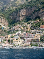 Colorful Mediterranean houses are stacked along a steep hillside overlooking a deep blue sea. The architecture features a blend of warm tones, with a prominent dome and tower standing out against the vibrant buildings. Lush greenery contrasts with the rugged cliffs in the background.