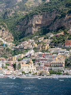 Colorful Mediterranean houses are stacked along a steep hillside overlooking a deep blue sea. The architecture features a blend of warm tones, with a prominent dome and tower standing out against the vibrant buildings. Lush greenery contrasts with the rugged cliffs in the background.