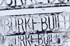 A close-up of a metal structure with the words 'BURKE BUILT' cut out, covered partially by snow. The lettering is clear against the white background.