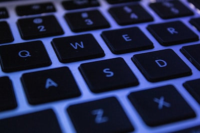 A close-up shot of a computer keyboard, highlighting several black keys with white letters on their surfaces. The lighting has a bluish tint, casting a cool tone on the keys. The image features keys like Q, W, E, R, A, S, D, and Z.