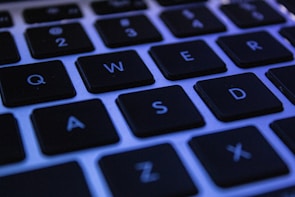 A close-up shot of a computer keyboard, highlighting several black keys with white letters on their surfaces. The lighting has a bluish tint, casting a cool tone on the keys. The image features keys like Q, W, E, R, A, S, D, and Z.